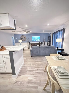 Una cocina moderna con gabinetes blancos, un comedor con mesa y sillas, un sofá gris y un televisor al fondo.