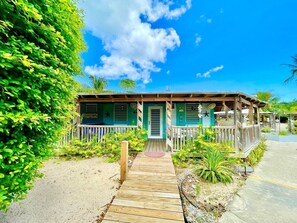 Une propriété située en bord de mer, dotée d’une terrasse en bois menant à une maison, le tout entouré d’une végétation luxuriante et surplombé d’un ciel bleu azur.