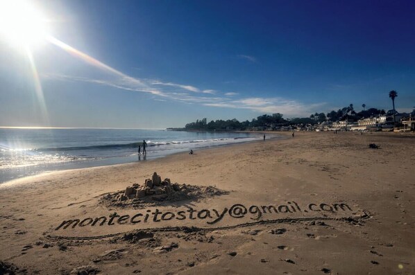 Une plage parsemée d’empreintes et sur laquelle on peut lire un message écrit dans le sable, un ciel dégagé et des bâtiments qui se dressent au loin.