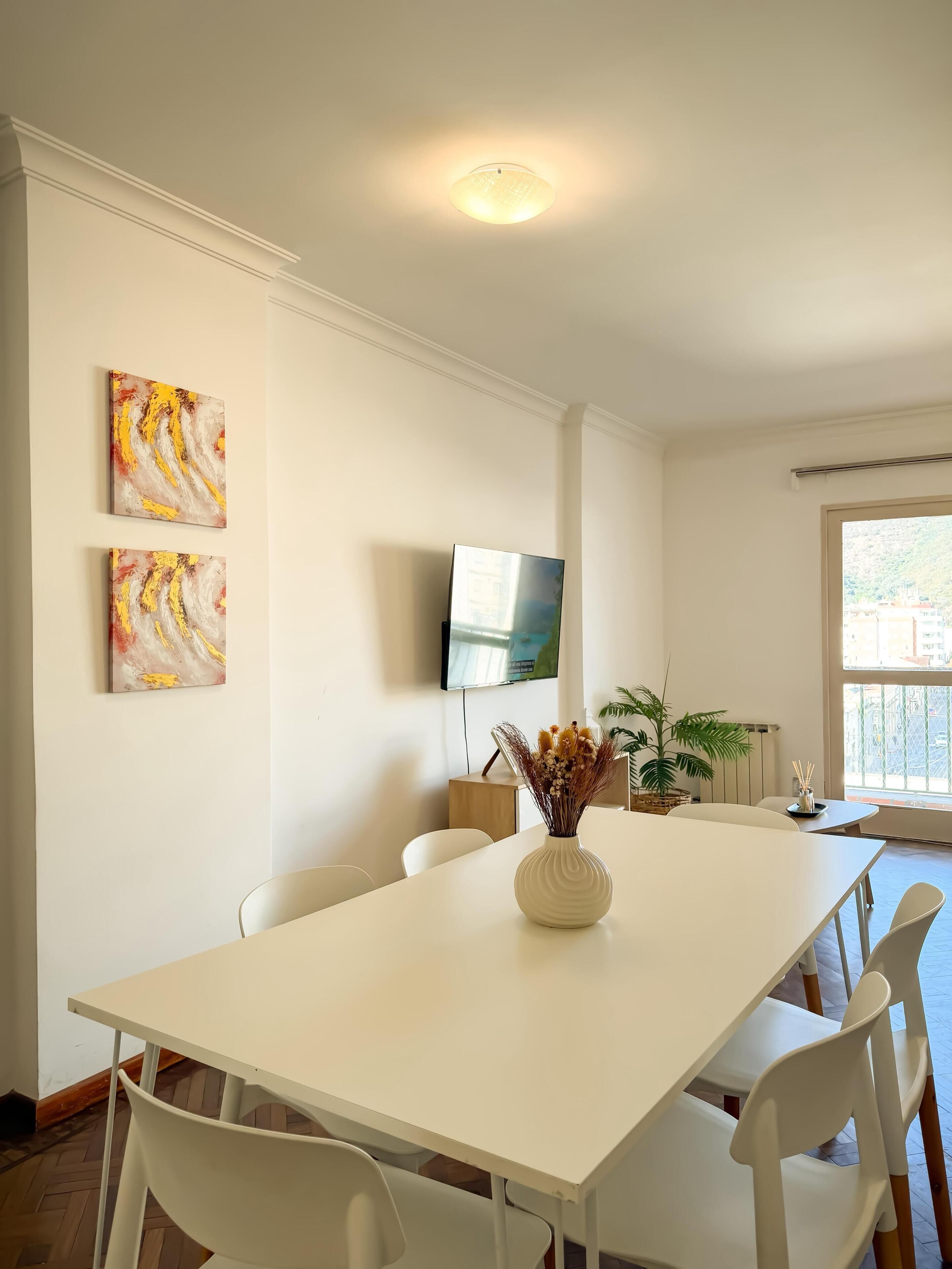 Un comedor con una mesa blanca, sillas blancas, dos cuadros abstractos, un televisor de pantalla plana, una planta en maceta y una ventana con vistas.