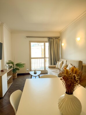 Ein modernes Wohnzimmer mit Sofa, Esstisch, Fernseher und einem Fenster mit Vorhängen.