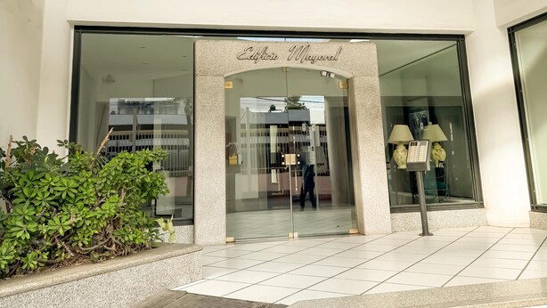 A modern building entrance with a glass door, a nameplate above the door, and a small garden area with plants. - Dpto en pleno centro de Salta con cochera (Salta)