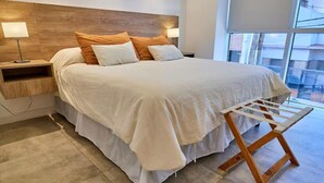 A modern bedroom with a large bed, wooden headboard, bedside tables, and a window with a view.
