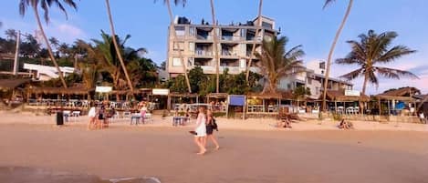 A beach with palm trees, a building with balconies, and people on the sand.