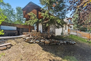 Ein modernes Haus mit Steinfundament, einer Holzterrasse und einem großen Baum, der Schatten spendet.