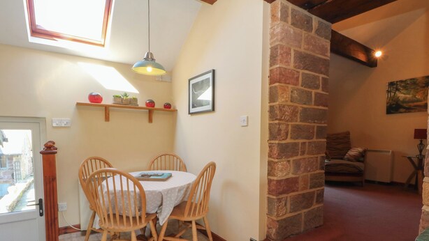 A dining area with a round table and four chairs, a hanging light fixture, a stone wall, and a painting on the wall. - Pearls Farm Two (Macclesfield)