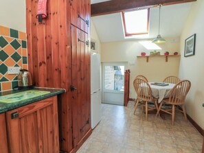 A kitchen with wooden cabinets, a refrigerator, a dining table with chairs, a window, and a ceiling light. - Pearls Farm Two (Macclesfield)