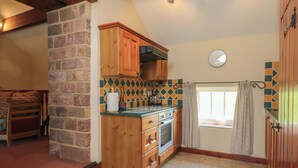 A kitchen with wooden cabinets, a stone wall, a tiled backsplash, a window with curtains, and a clock on the wall. - Pearls Farm Two (Macclesfield)