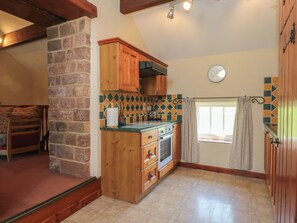 A kitchen with wooden cabinets, a stone wall, a tiled backsplash, a window with curtains, and a clock on the wall. - Pearls Farm Two (Macclesfield)