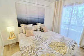Ein Schlafzimmer mit einem Bett, einem Nachttisch, einer Lampe, einem großen Gemälde an der Wand und einem Fenster mit Vorhängen.