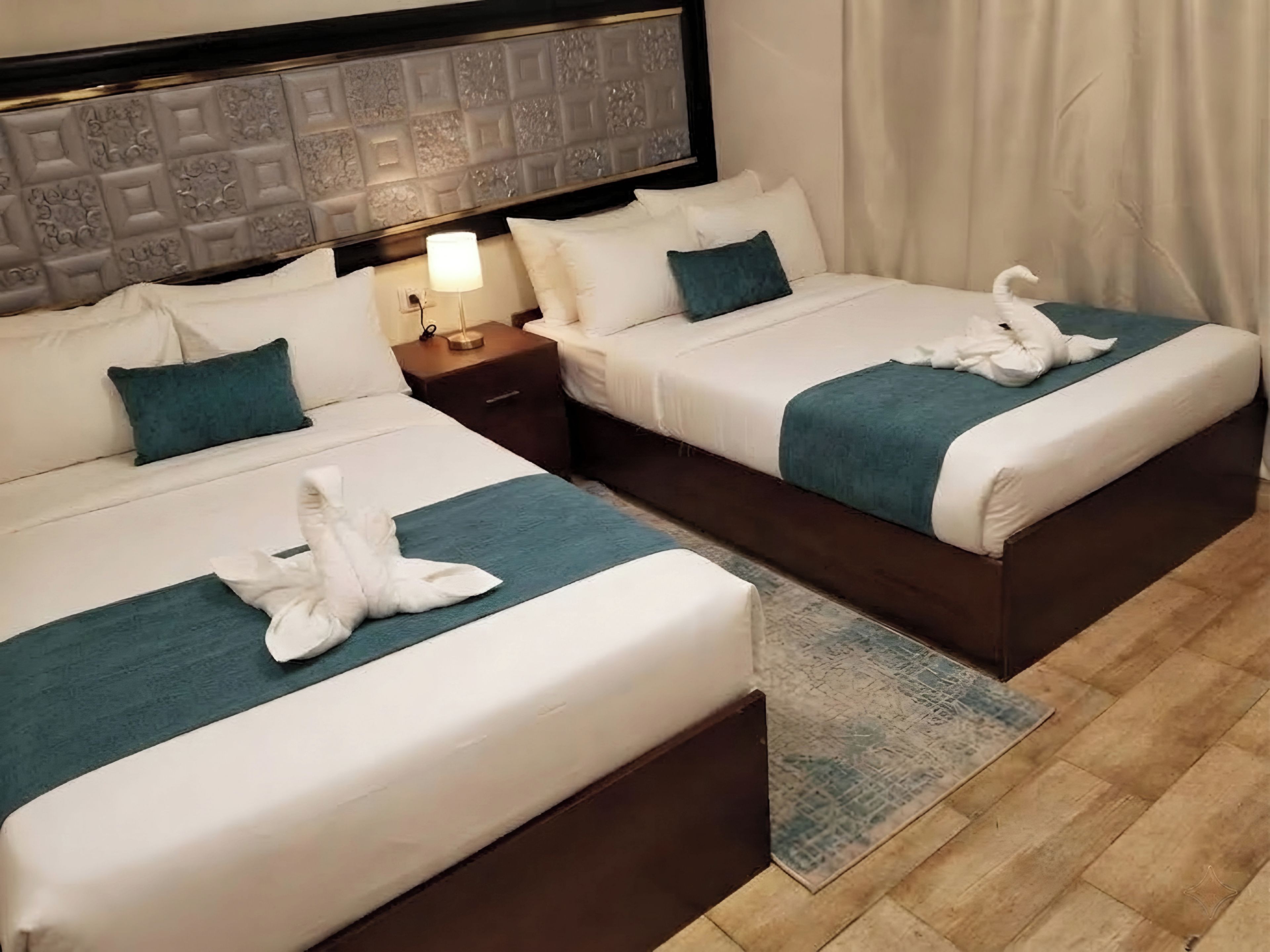 A hotel room with two beds, each with a white duvet and teal throw pillows, a nightstand with a lamp, and a decorative headboard.