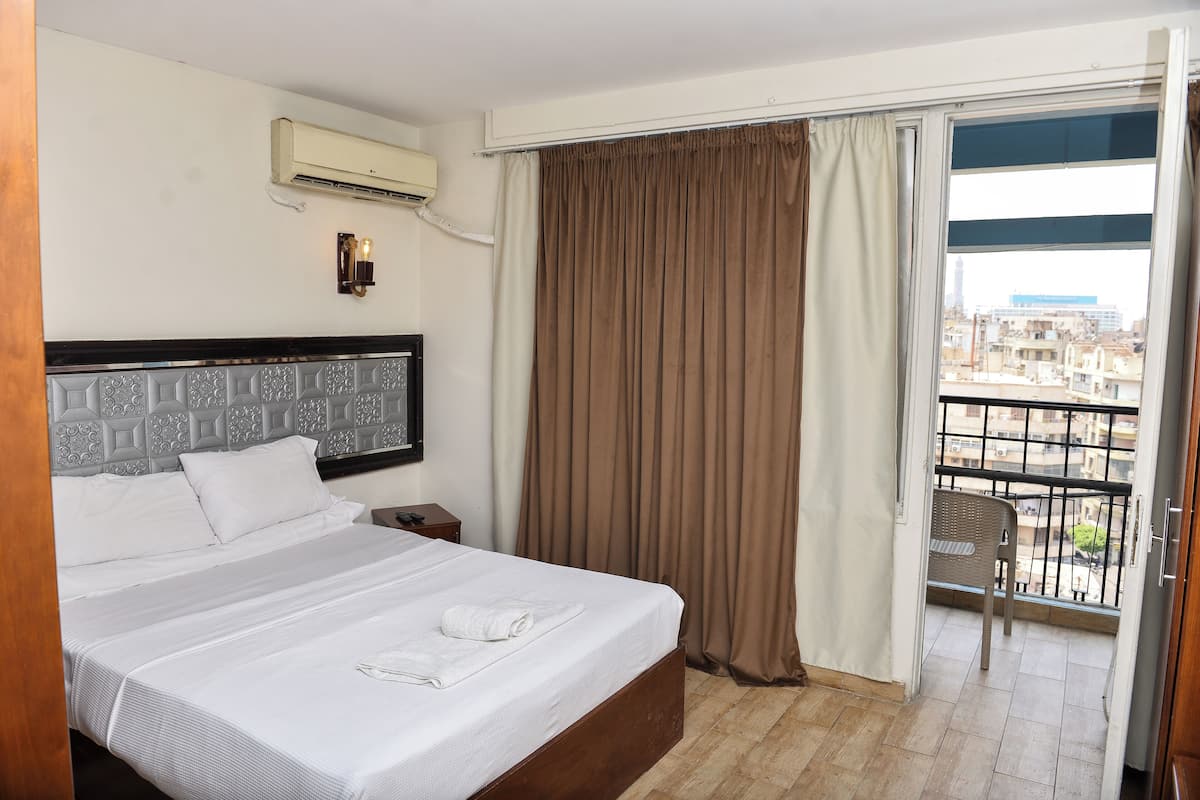 Quarto de hotel com uma cama grande, uma mesa de cabeceira, ar condicionado, uma janela com cortinas e vista para a cidade.