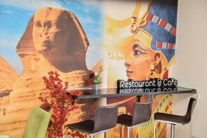 Mostrador de restaurante con un mural de la Esfinge y una mujer con traje tradicional, dos sillas y un jarrón con flores.