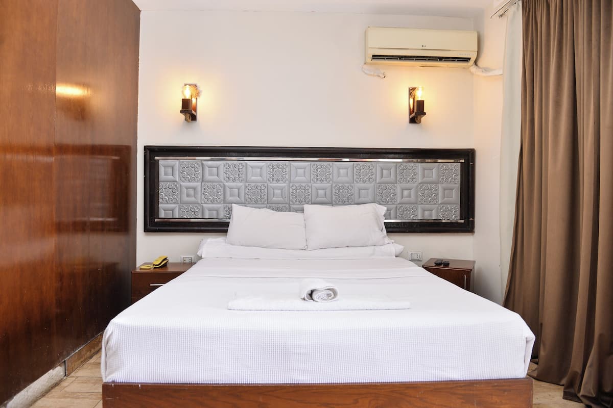 Uma cama bem arrumada com colcha e almofadas brancas, cabeceira com padrão geométrico e duas luminárias fixadas na parede.