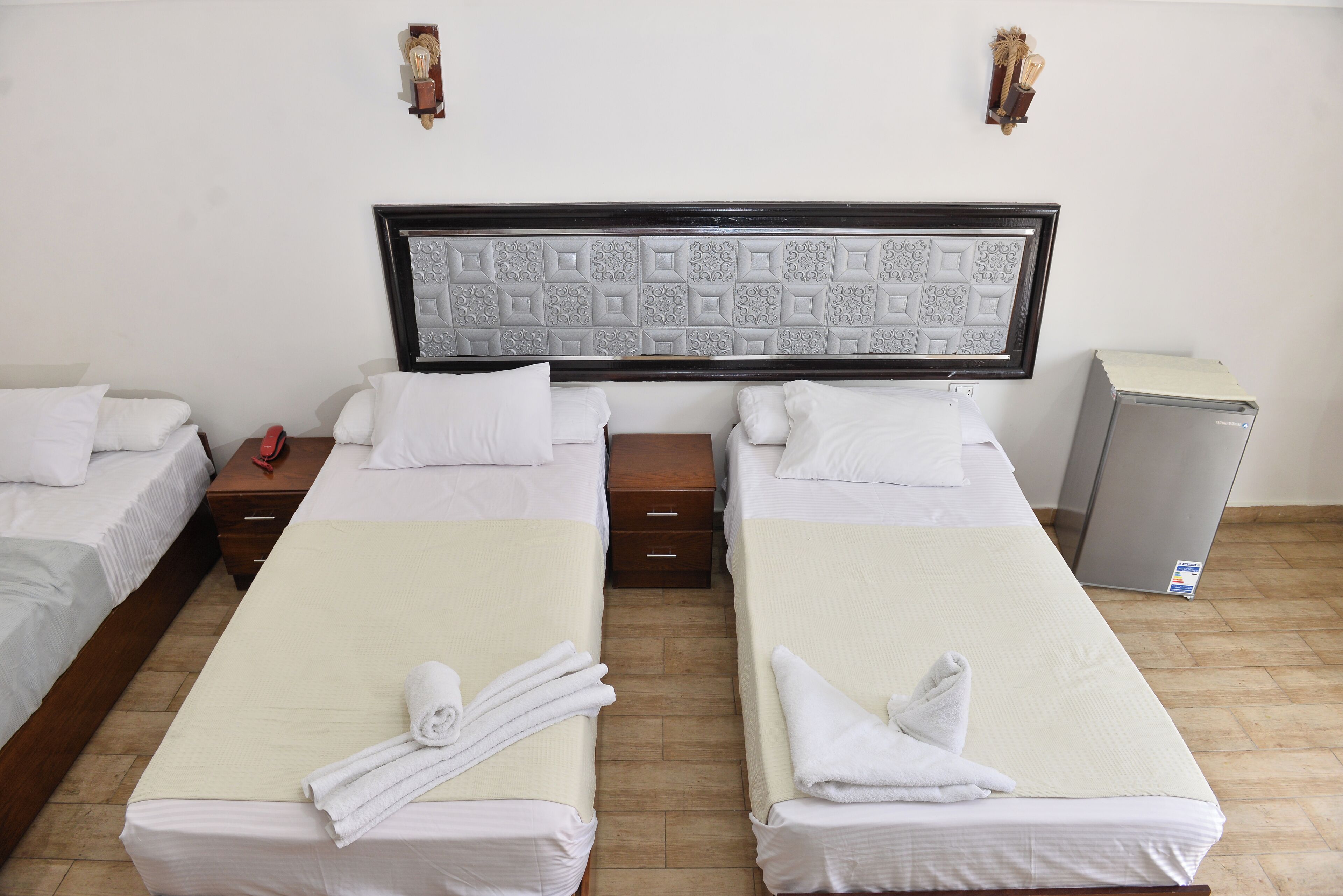 A hotel room with two single beds, a wooden nightstand, a small table, a wall-mounted light fixture, and a compact refrigerator.