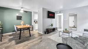 Uma sala de estar moderna com uma televisão de tela plana, uma área de jantar com mesa e cadeiras de madeira e um sofá cinza.