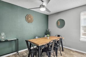 Un comedor con mesa de madera, sillas negras, ventilador de techo y decoración en la pared.