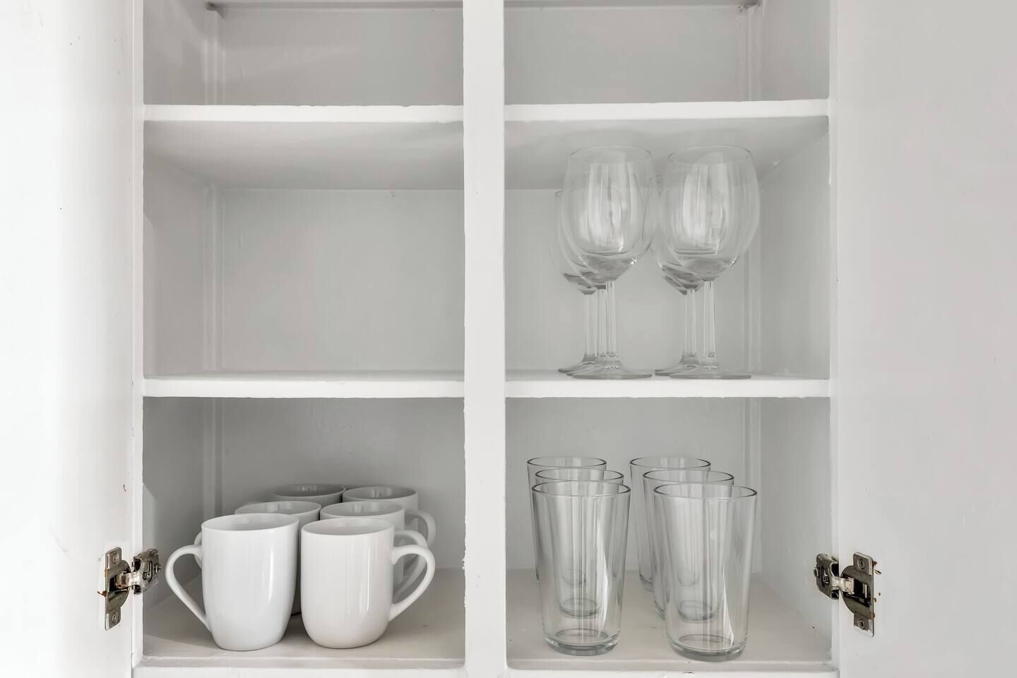 Un meuble blanc avec plusieurs étagères. L’étagère supérieure contient deux verres à vin transparents. L’étagère du milieu contient plusieurs tasses blanches. L’étagère inférieure contient plusieurs verres transparents.