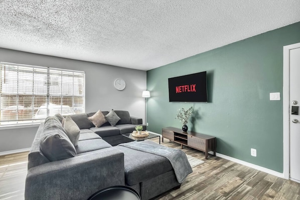 Una sala de estar moderna con un sofá modular, un televisor montado en la pared y un ventanal con persianas.