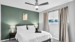 A bedroom with a bed, a nightstand, a ceiling fan, a window with curtains, and a wall with a single framed picture.