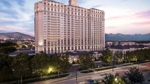 A large, multi-story hotel with a prominent tower, surrounded by trees and a road with moving vehicles. - 2 Queen Suite | Grand America | Grand Spa, pools, wellness classes (Salt Lake City)