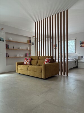 Un salon moderne avec un canapé, des étagères, un élément de séparation décoratif et un lit à l’arrière-plan.
