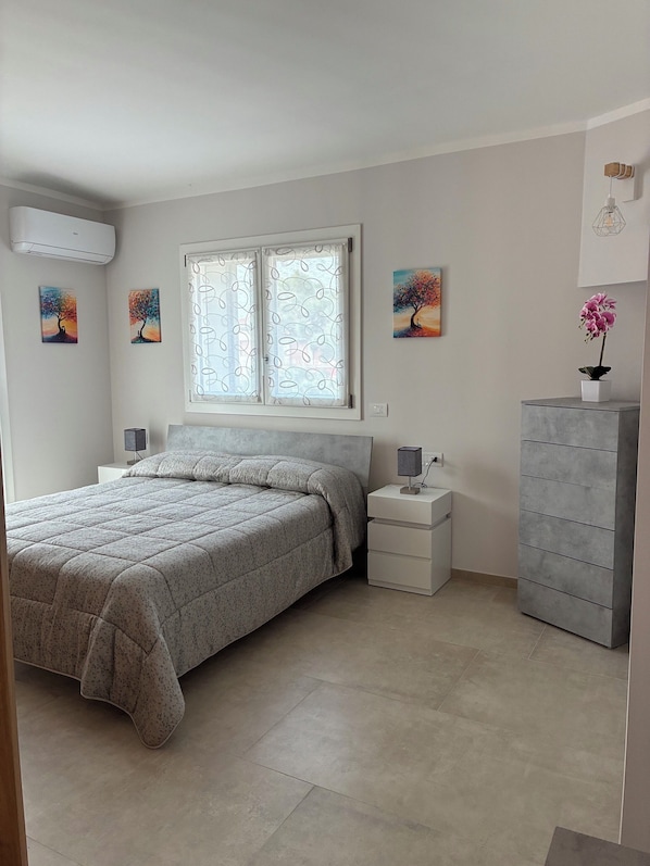 Une chambre à coucher comprenant un lit, une table de chevet, une commode, une fenêtre avec des rideaux et deux tableaux accrochés au mur.