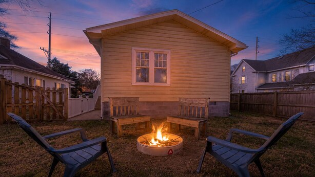 Ein Garten mit einem Feuerkorb, zwei Holzbanken und einem Haus mit Satteldach.