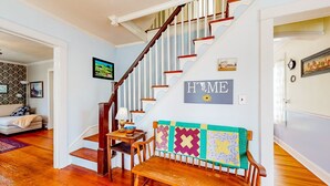 Eine Treppe mit Holzbalken und weißen Spindeln, eine Bank mit bunter Steppdecke und ein 'HOME'-Schild an der Wand.