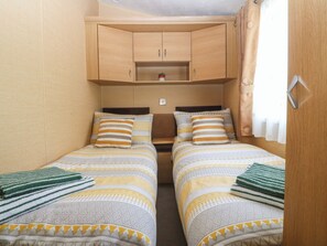 A small, compact room with two beds, wooden cabinets above, and a window with curtains. - Temperance Caravan (Holyhead)