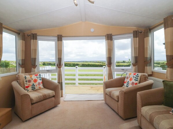 A room with two sofas facing a large window, a wooden floor, and a view of a white fence and green fields. - Temperance Caravan (Holyhead)