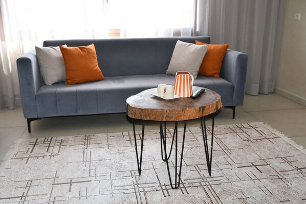 Un salon moderne avec un canapé gris, des coussins orange et blancs, une table basse ronde sur laquelle se trouve une tasse à motifs et un tapis à motifs.