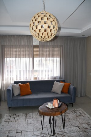 Un salon avec un canapé bleu, une table basse ronde en bois et un luminaire suspendu en fibres tressées.
