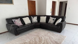 A L-shaped black leather sofa with white and patterned cushions, a beige patterned rug, and a white tiled floor. - Crest Premiums Sandton (Johannesburg)