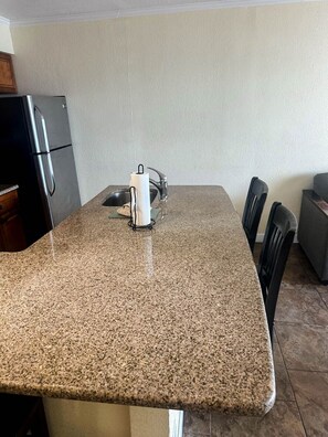 A kitchen with granite countertops, a stainless steel refrigerator, a sink, and a dining area with chairs. - Wake Up to Stunning Views – Your Balcony Escape! (Tampa)