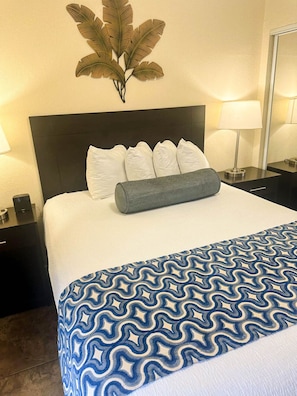 A hotel room with a large bed, a dark headboard, white pillows, a blue patterned bedspread, a wall-mounted plant, and a nightstand with a lamp. - Wake Up to Stunning Views – Your Balcony Escape! (Tampa)