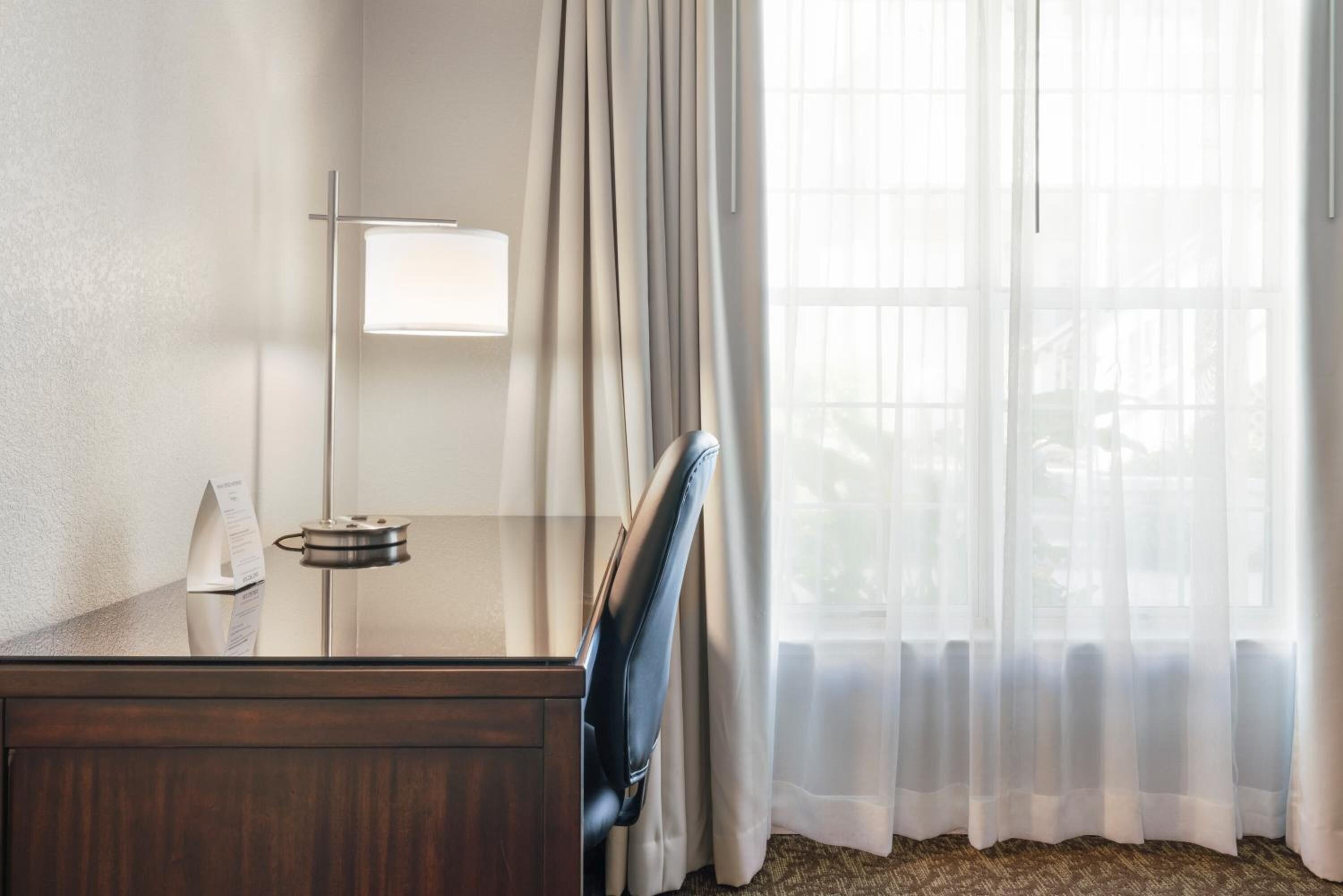 Habitación de hotel con escritorio de madera, lámpara, silla y cortinas traslúcidas.