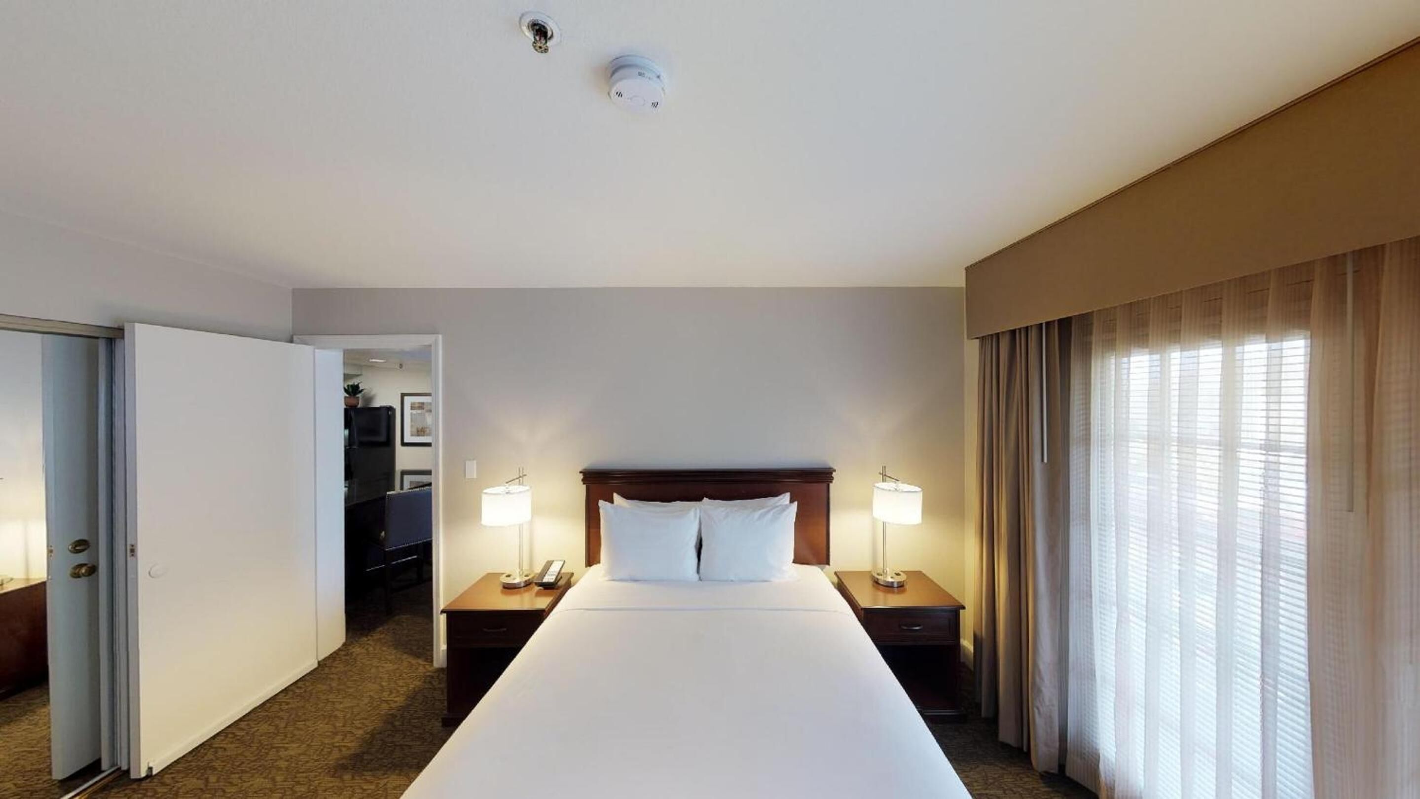 A hotel room with a large bed, two bedside tables, a desk, a lamp, and a window with curtains.