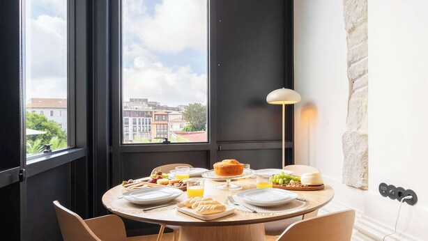 Mesa de jantar com itens de café da manhã, um abajur e vista para edifícios através de grandes janelas.