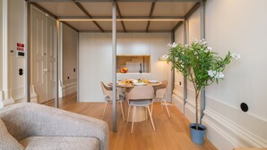Et moderne spiseområde med et rundt bord, stole, en potteplante og et synligt køkken i baggrunden.