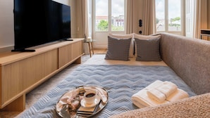 Et moderne hotelværelse med et stort fladskærms-tv, en seng med et blåt og gråt mønstret sengetæppe, et natbord med et bakke med morgenmad, og et vindue med udsigt over byen.