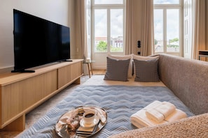 Ein modernes Hotelzimmer mit einem großen Flachbildfernseher, einem Bett mit einem blau-grauen Muster, einem Nachttisch mit einem Frühstücksangebot und einem Fenster mit Blick auf die Stadt.