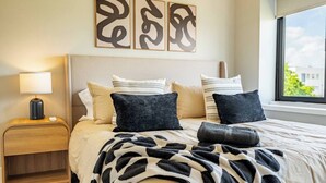 Une chambre à coucher avec un lit, des oreillers, une table de chevet, une lampe et des œuvres d’art abstraites accrochées au mur.