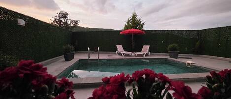 A swimming pool with two lounge chairs and a red umbrella, surrounded by a hedge and potted plants.
