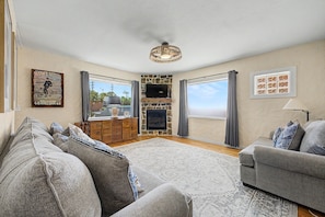 A living room with a stone fireplace, a television mounted on the wall, and a large window with curtains. - Midtown Tucson Casita | Near UofA & Dining (Tucson)