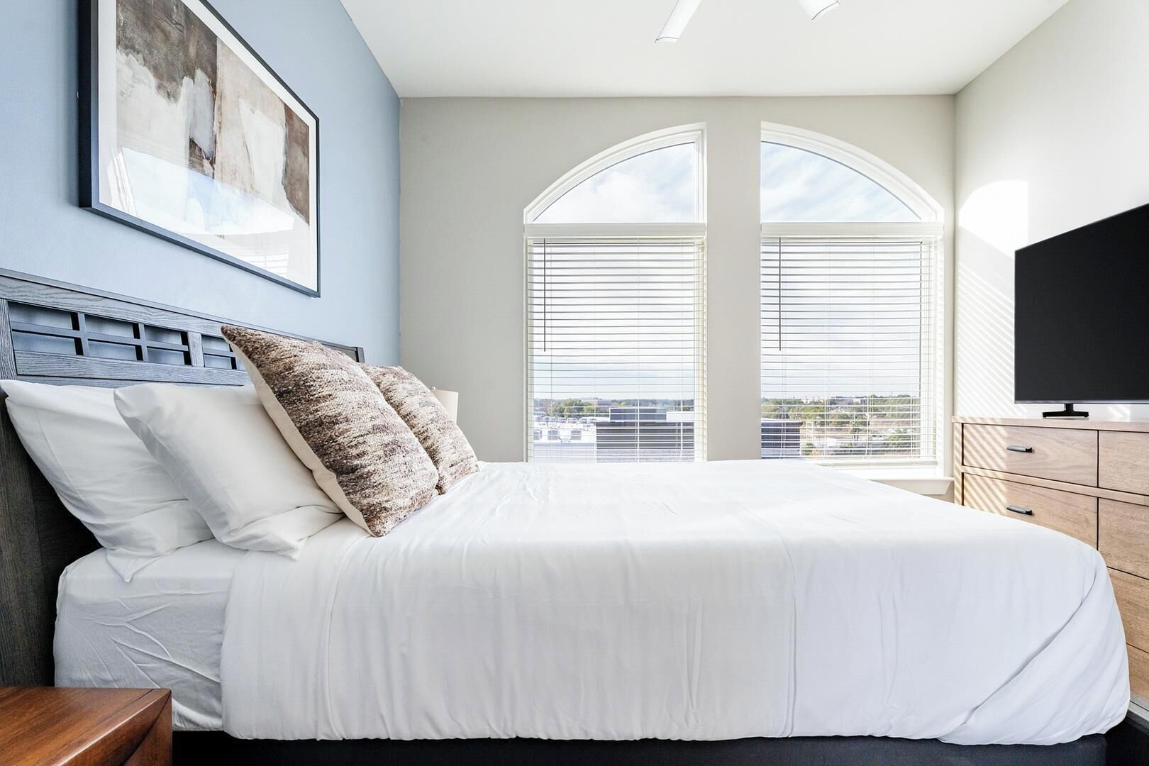A bedroom with a large bed, a TV, and a view of the city through the windows.
