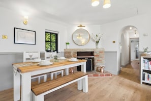 Un comedor con mesa de madera, bancos blancos, chimenea y estantería.