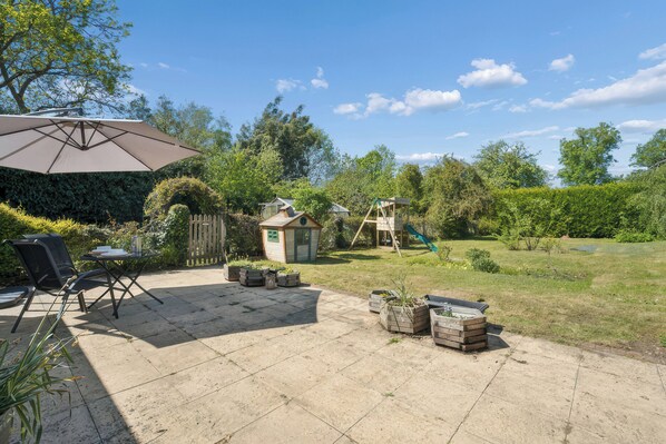 A patio with a table, chairs, and an umbrella. There is a small wooden structure and a swing set in the background.