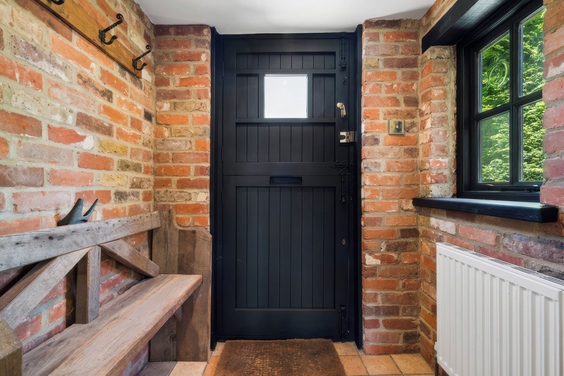 A black door with a small window above it, set in a brick wall with a wooden bench to the left and a window to the right.