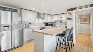 A modern kitchen with stainless steel appliances, white cabinetry, and a wooden island with bar stools. - 6-Bedroom Family Haven with Garden in Aylesbury (Aylesbury)